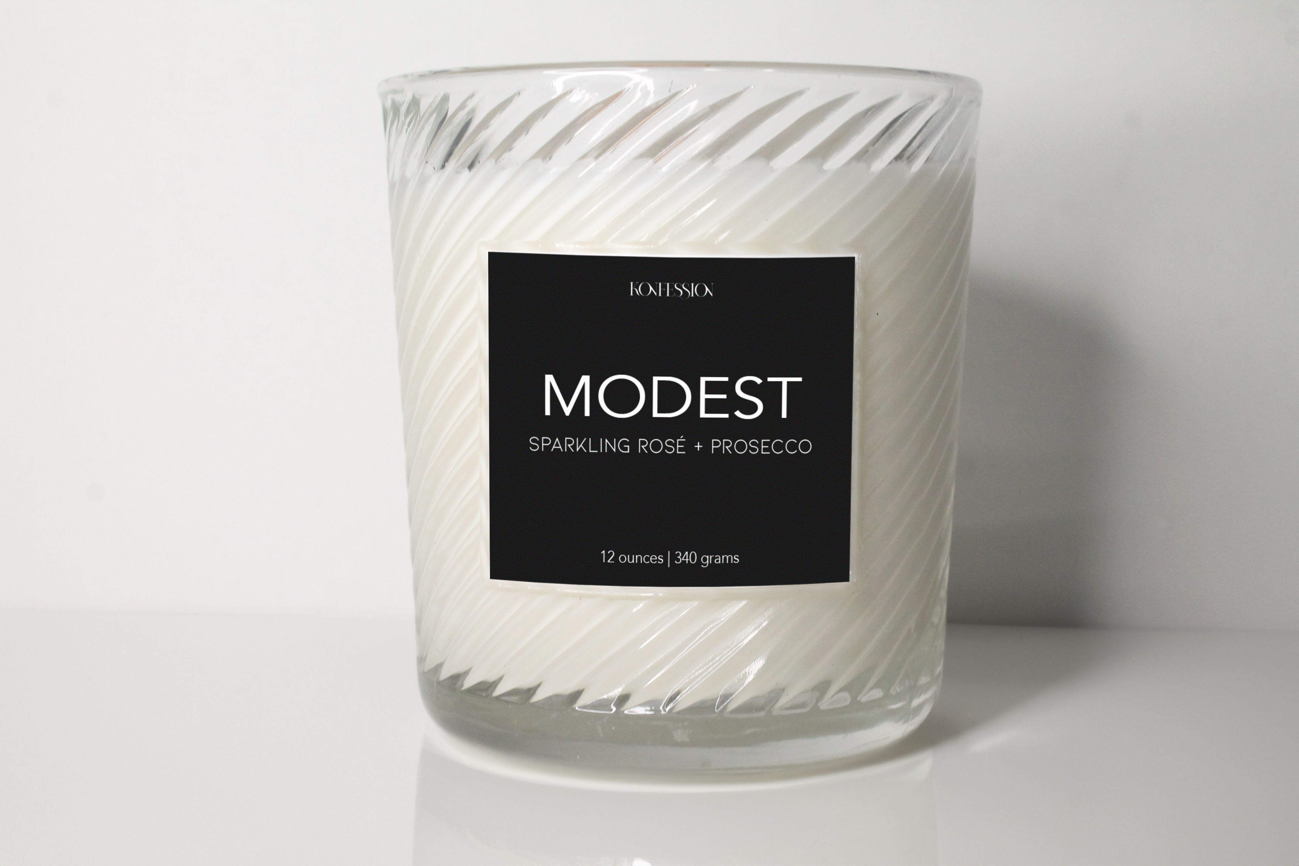 12 ounce soy wax candle. The candle is called Modest and it is a sparkling rose and prosecco scent. It has a black label on ribbed clear glass vessel.