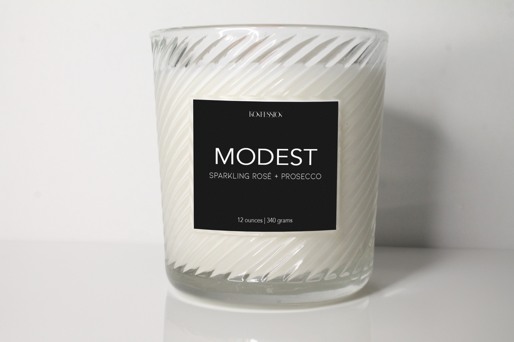12 ounce soy wax candle. The candle is called Modest and it is a sparkling rose and prosecco scent. It has a black label on ribbed clear glass vessel.